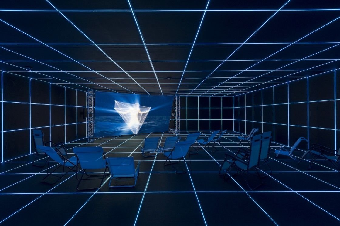 Hito Steyerl, Factory of the Sun: phòng chiếu ánh xanh với lưới đường sáng trên tường và sàn, ghế nằm bãi biển trước màn hình