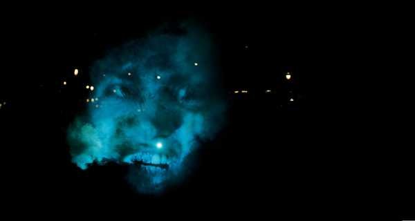 Tony Oursler, The Influence Machine: khuôn mặt chiếu lên làn khói và tán cây trong công viên ban đêm